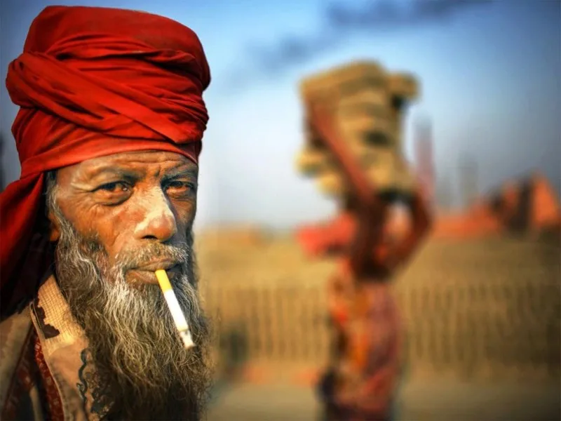 A weathered Bangladeshi man wearing a red turban smokes a cigarette, his expressive face capturing the raw character of rural life.