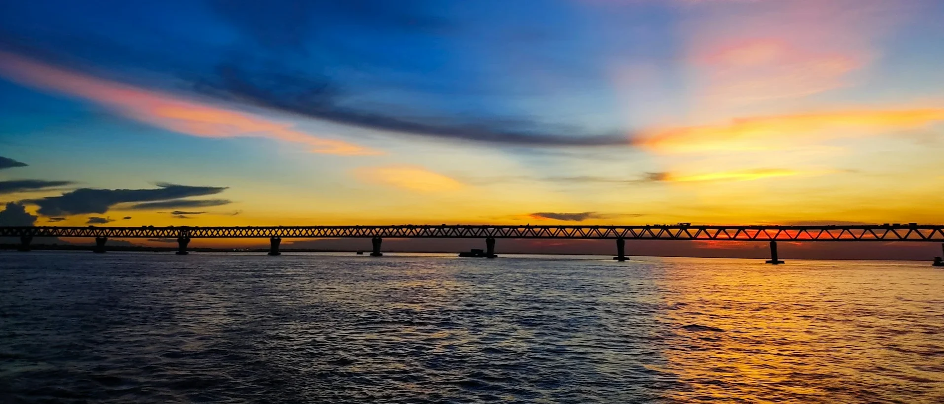 A wide-angle silhouette of the Padma Bridge stretching across the river under a vibrant sunset sky with hues of blue, orange, and pink.