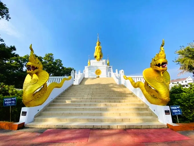 Beautiful blue sky and green trees next to the white stairs, toward a golden Buddha statue, flanked by two large golden Naga serpent sculptures.