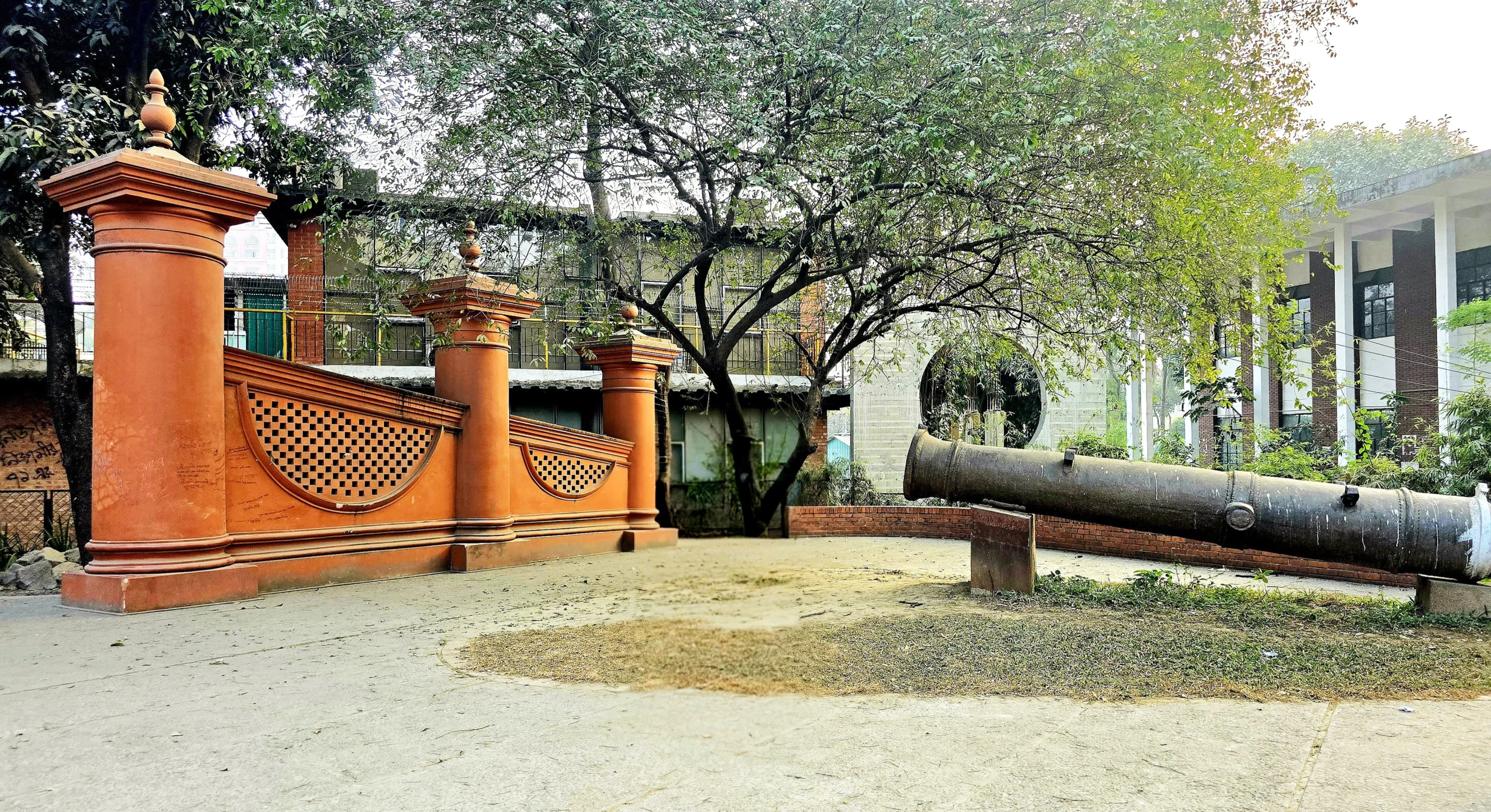 The renovated Dhaka Gate featuring three yellow-orange pillars and a historic Mughal-era cannon on display in a landscaped plaza near Dhaka University.