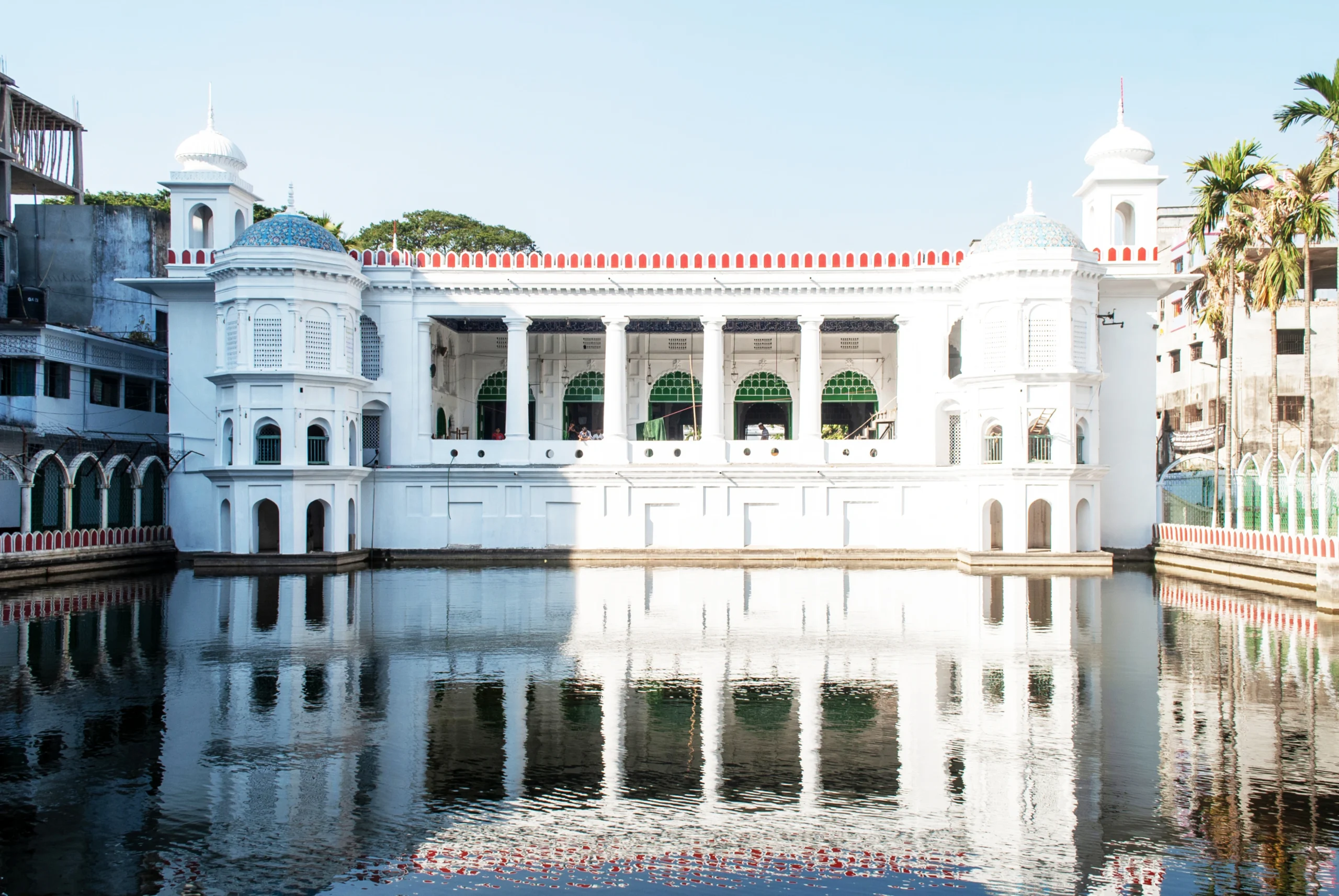 The white facade of Hussaini Dalan reflecting in a large still pond, showcasing its blend of Mughal and British architectural styles under a bright sky.