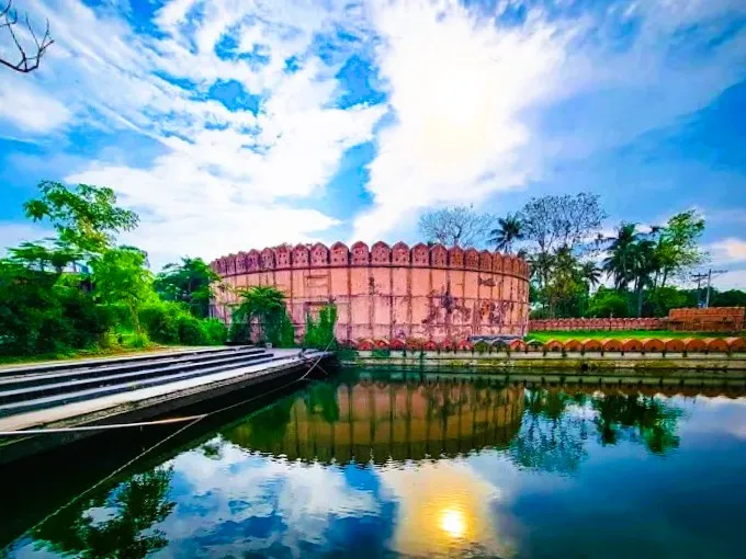 The circular defensive wall of Idrakpur Fort reflecting in the water, surrounded by lush green trees under a bright blue sky with a bird flying overhead.