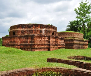 Three large, circular red brick foundations of ancient Buddhist stupas at Kutila Mura, set on a grassy hill under a cloudy sky.