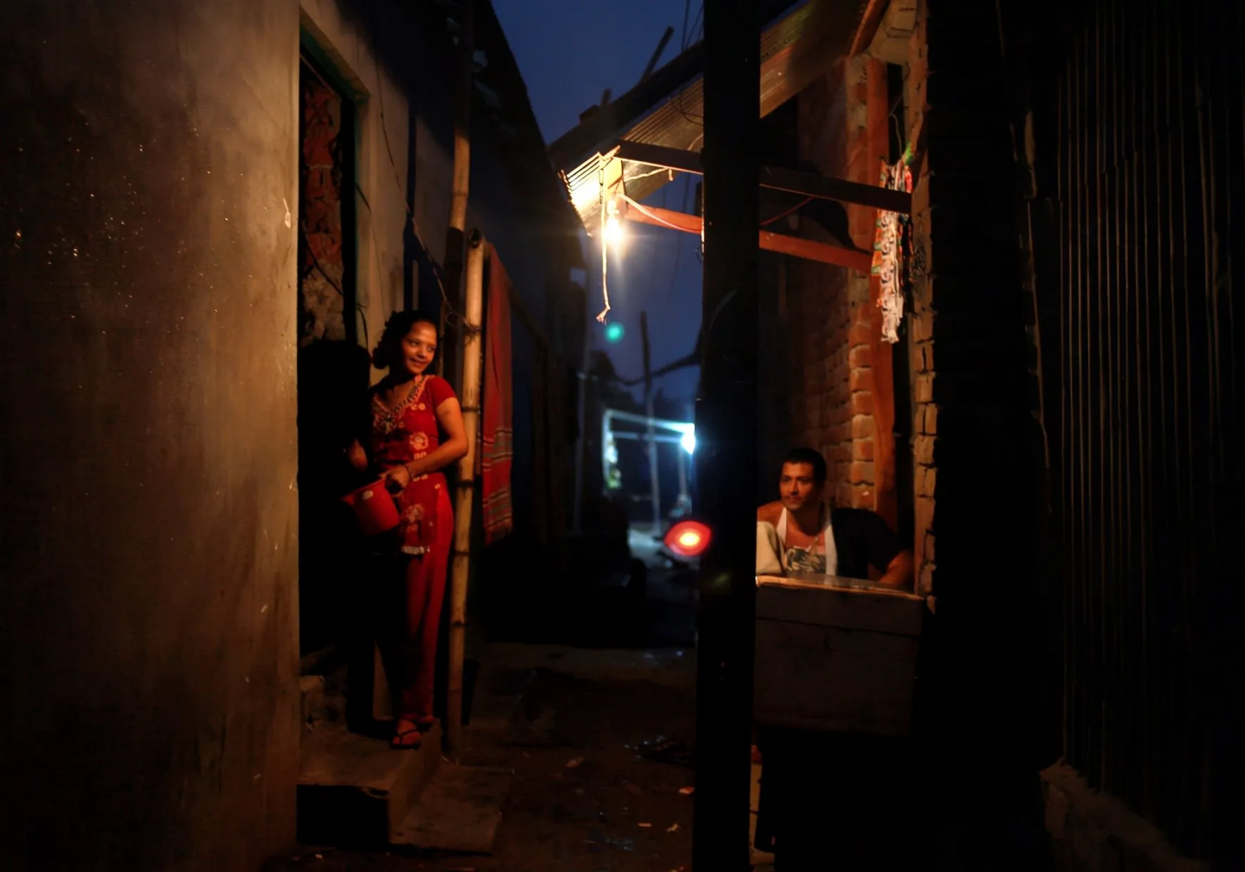 A day in the life of sex workers in Bangladesh showing a woman in a red outfit standing under a low hanging light bulb in a dark alleyway at night.